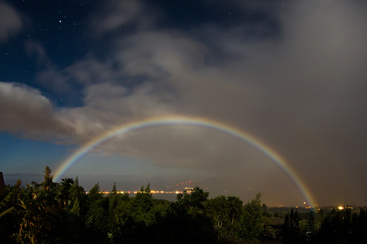 A pale lunar rainbow arcing through mist under a moonlit night sky